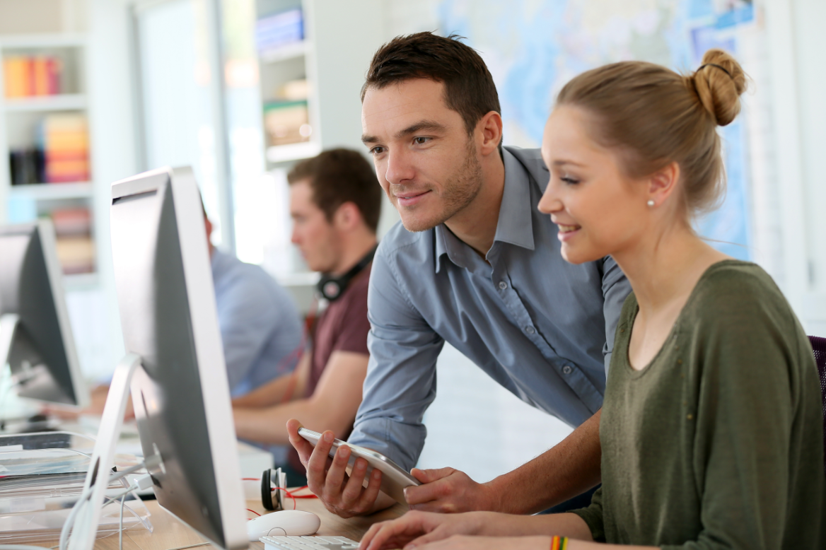 A manager training an employee, both looking at a desktop computer monitor.