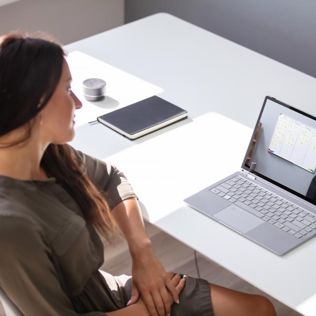 A woman sits at a desk and watches a video on her laptop of a lecturer pointing at a whiteboard.