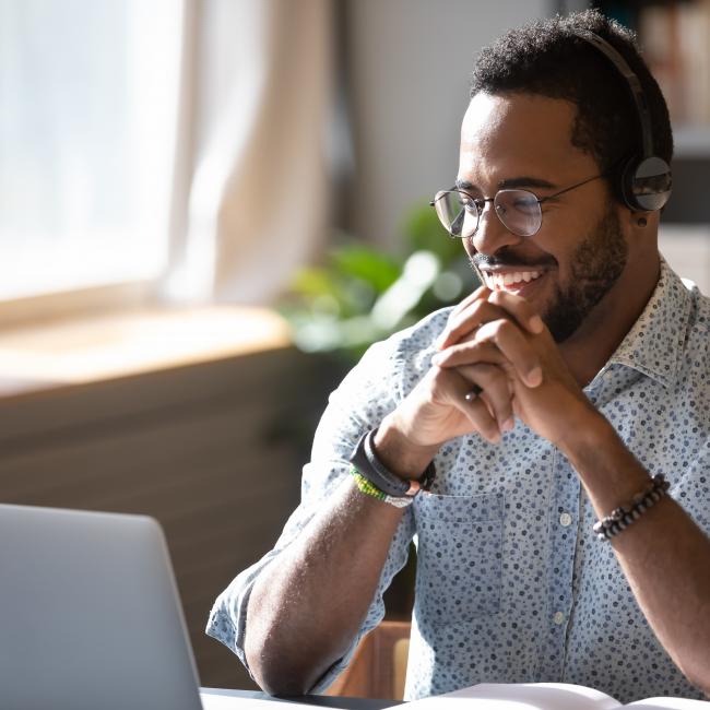 Man with headphones sitting at a desk, laughing at laptop monitor