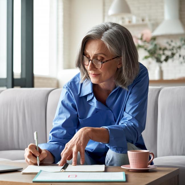 A woman writing in a notebook on a table, with an open laptop facing her on the table 