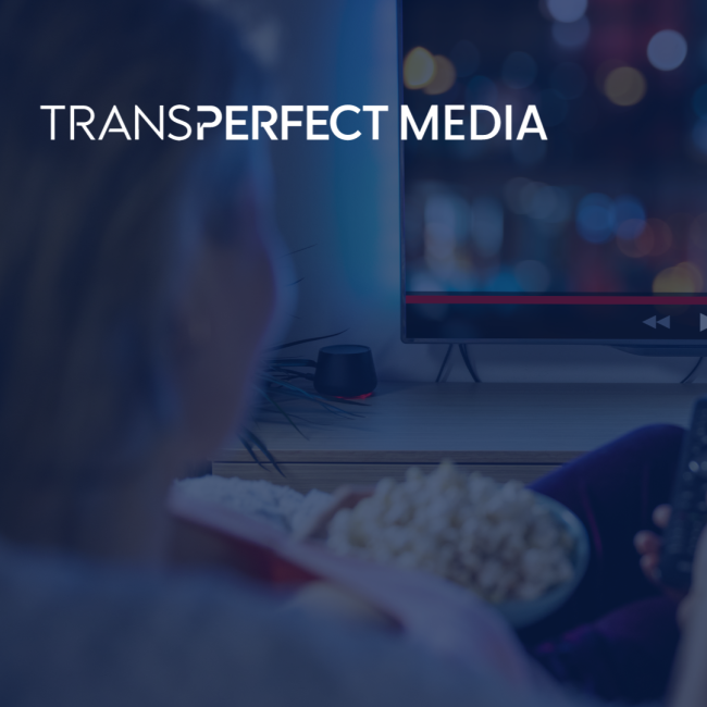 A woman sits on her couch with popcorn and a tv remote pointed at her tv while she pauses/plays a film playing. "TransPerfect Media" logo in the corner of image.