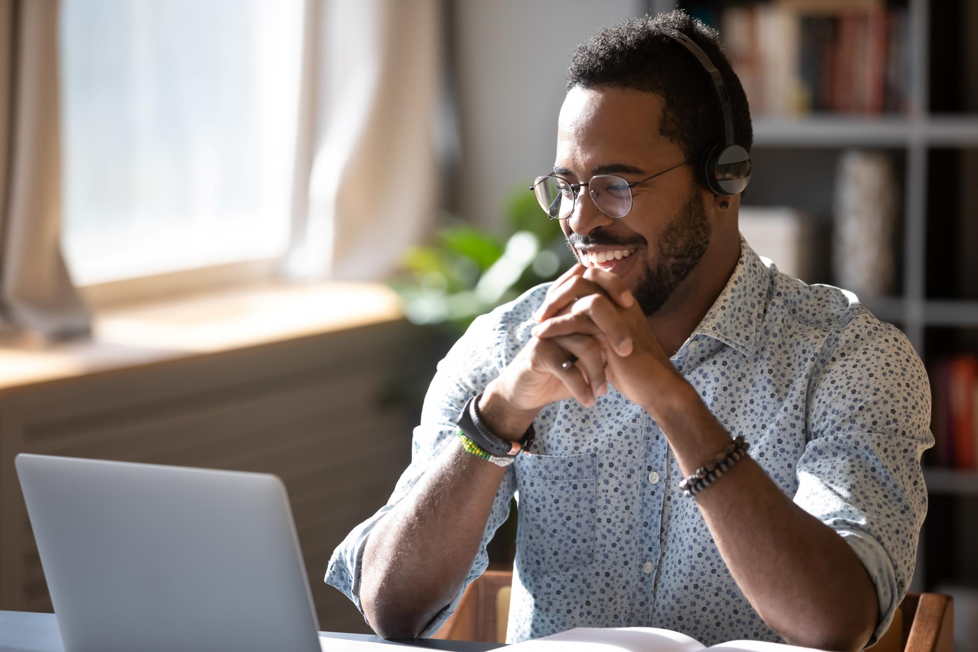 Man with headphones sitting at a desk, laughing at laptop monitor