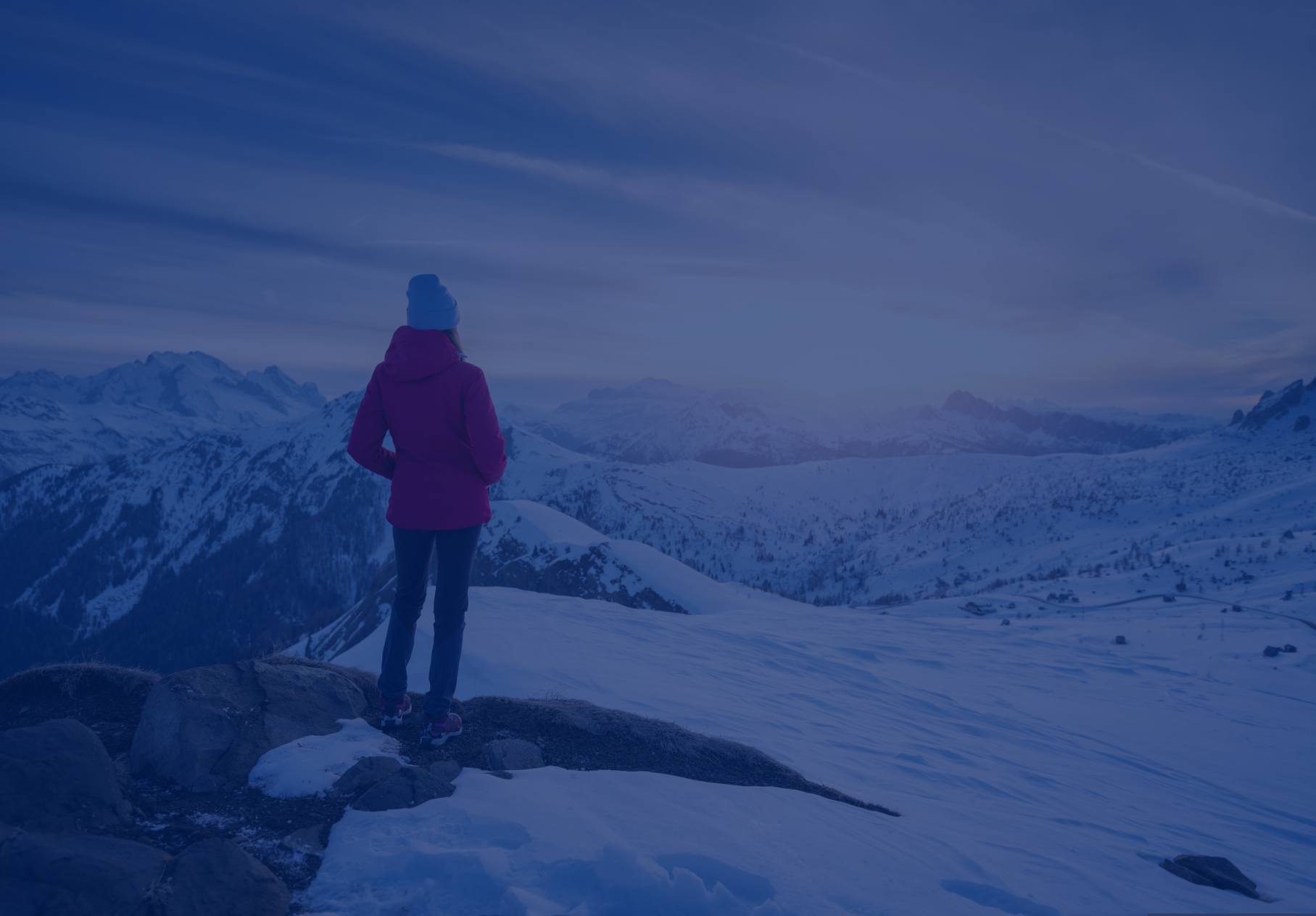 A person in stands on a hilly landscape overlooking snowy mountains.